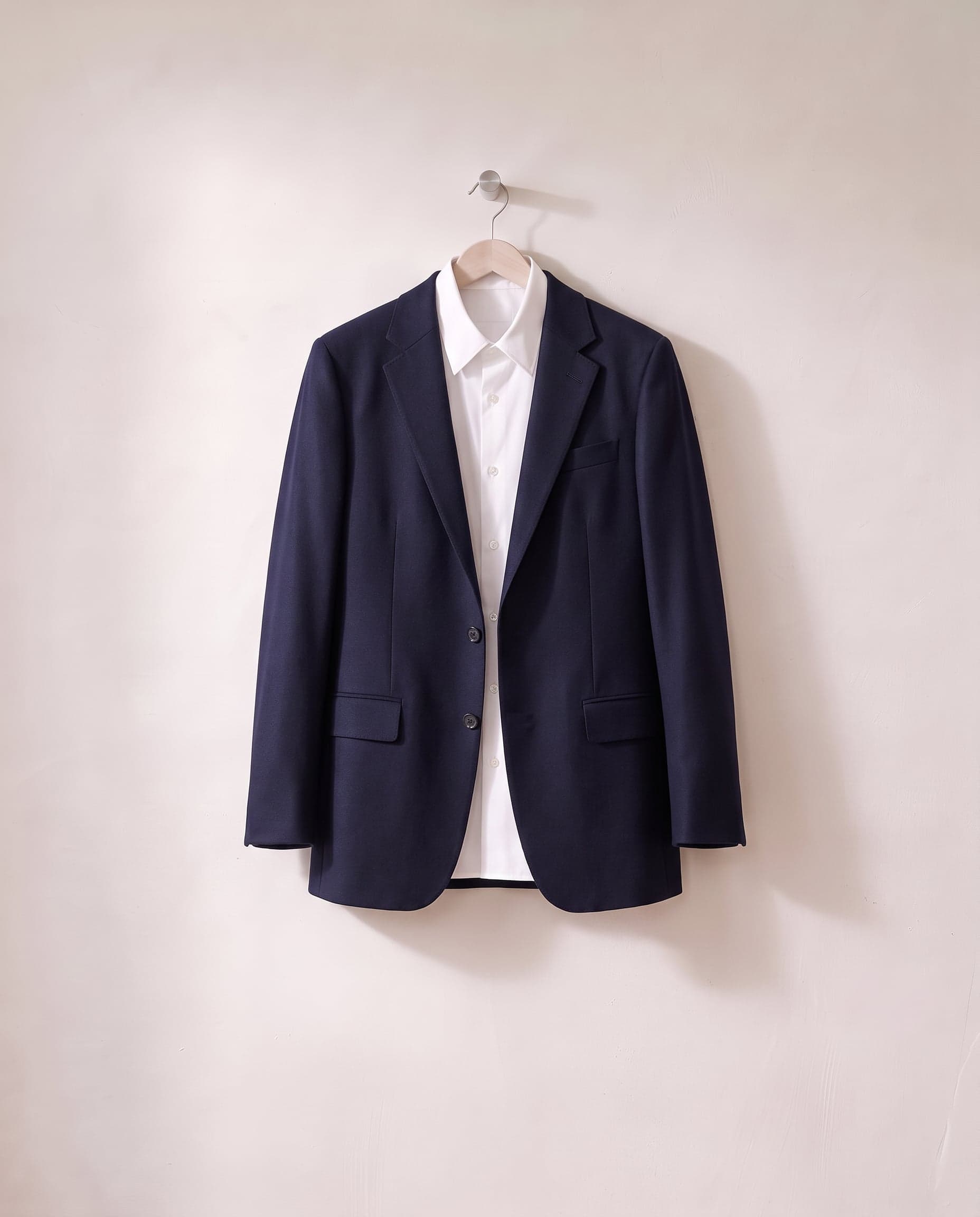 A navy tailored jacket over a white shirt on one wooden hanger, against a white plaster wall.