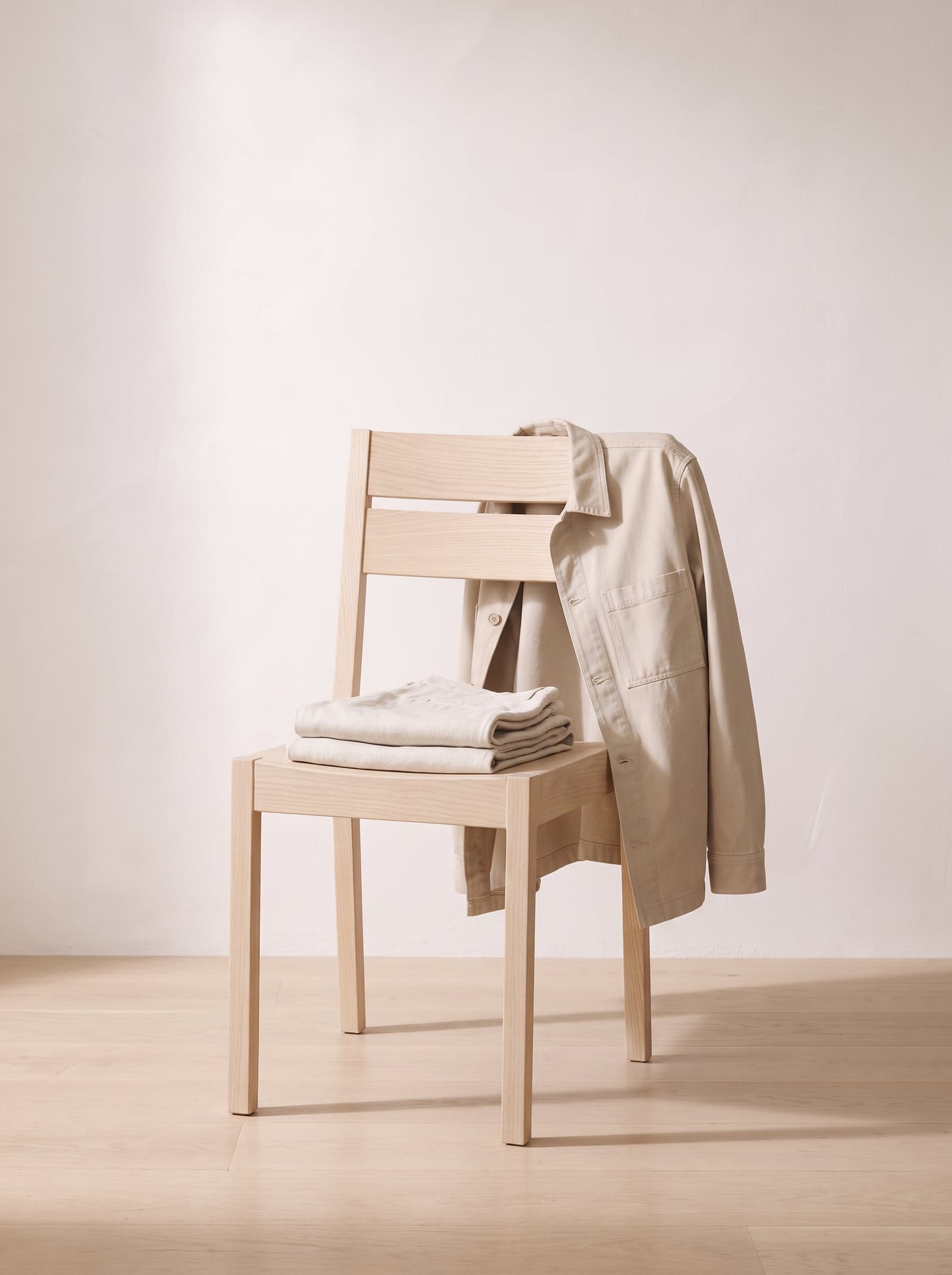 A stone overshirt over the back of a wooden chair, with folded linen trousers on the seat.