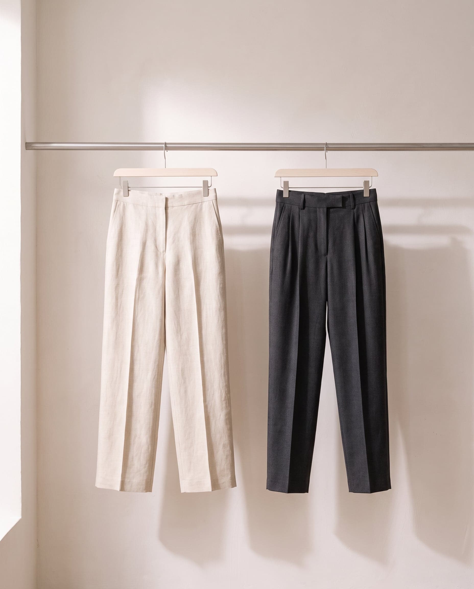 Linen and charcoal wool trousers hanging side by side from clip hangers on a steel rail.