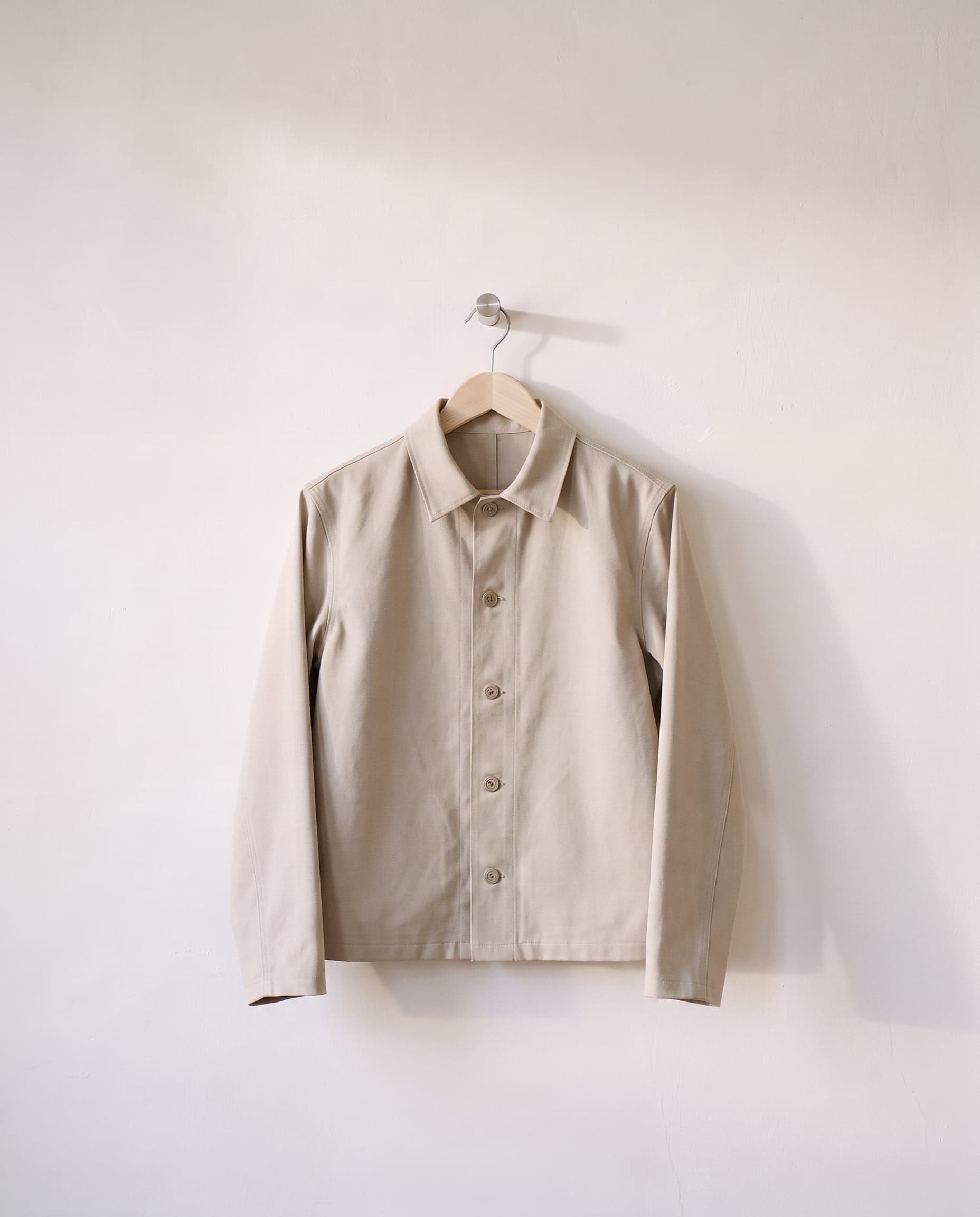 A finished stone cotton twill jacket with tonal buttons on a wooden hanger, against a white plaster wall.