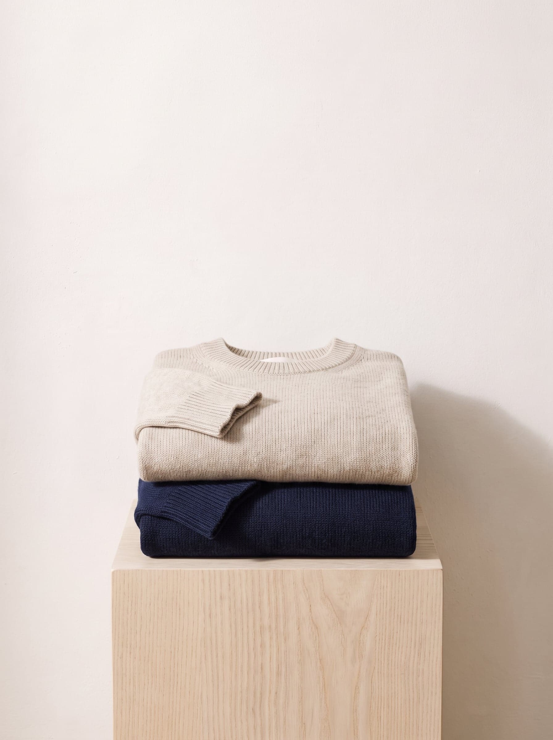 Two folded crewneck sweaters, oatmeal and navy, on a low wooden block.
