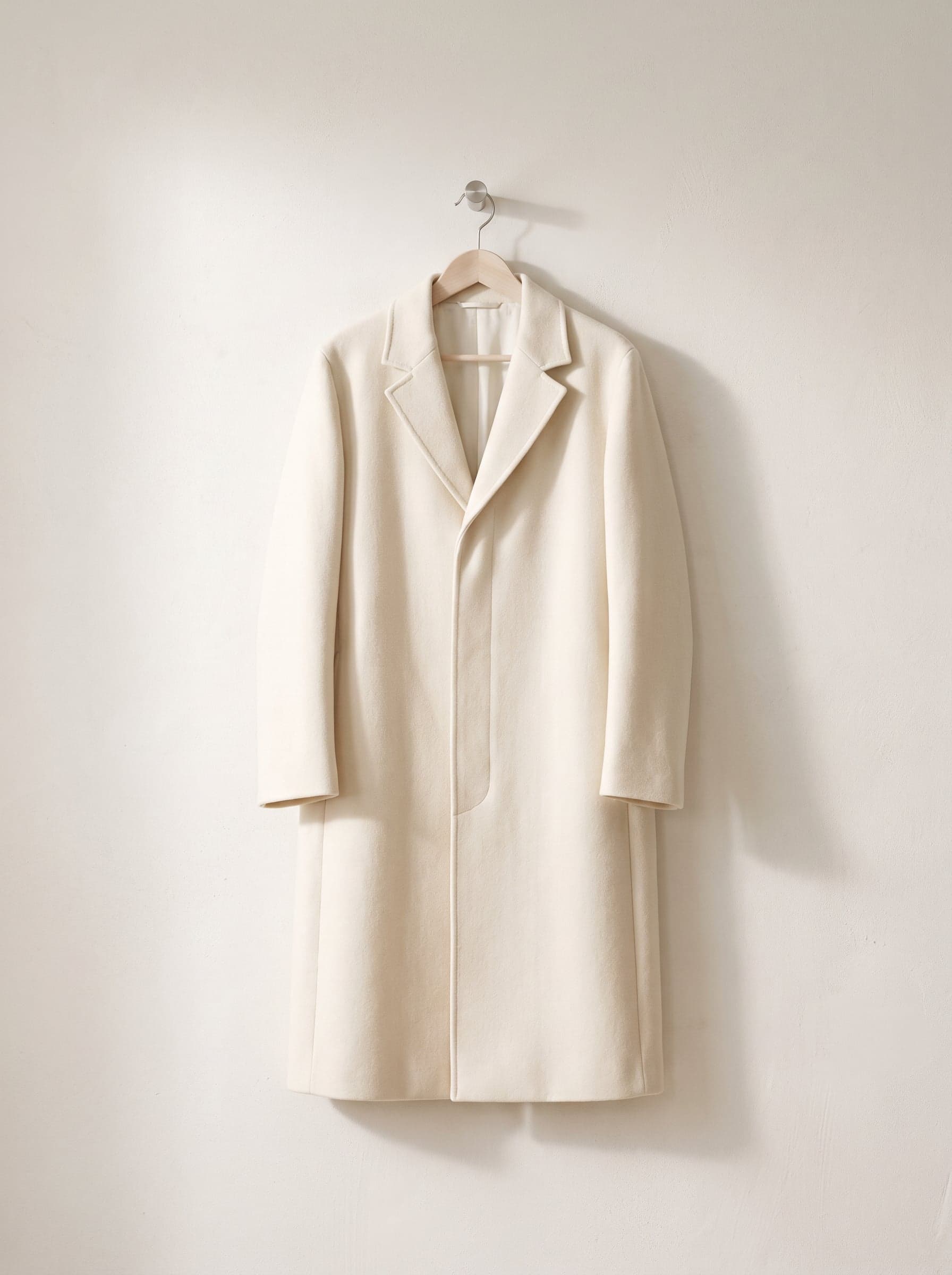Ecru long coat on a wooden hanger against a white plaster wall.