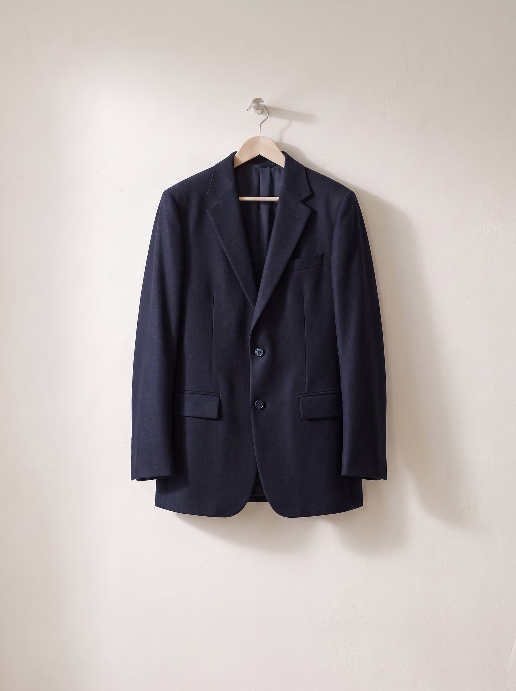 Navy tailored jacket on a wooden hanger against a white plaster wall.