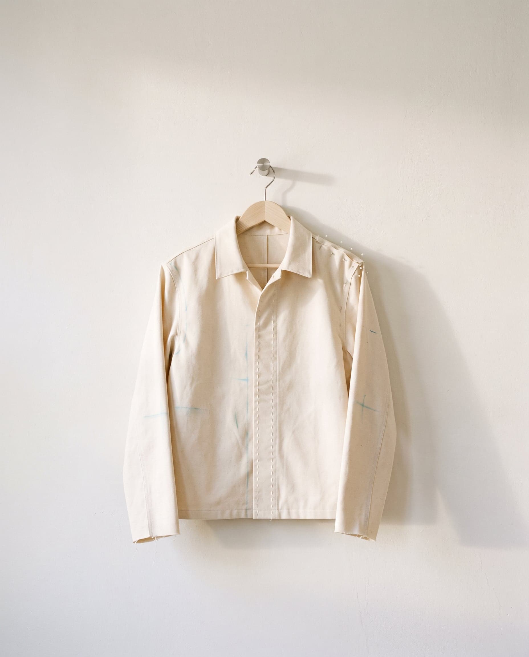 An unfinished calico sample jacket with basting stitches and chalk marks, on a hanger.