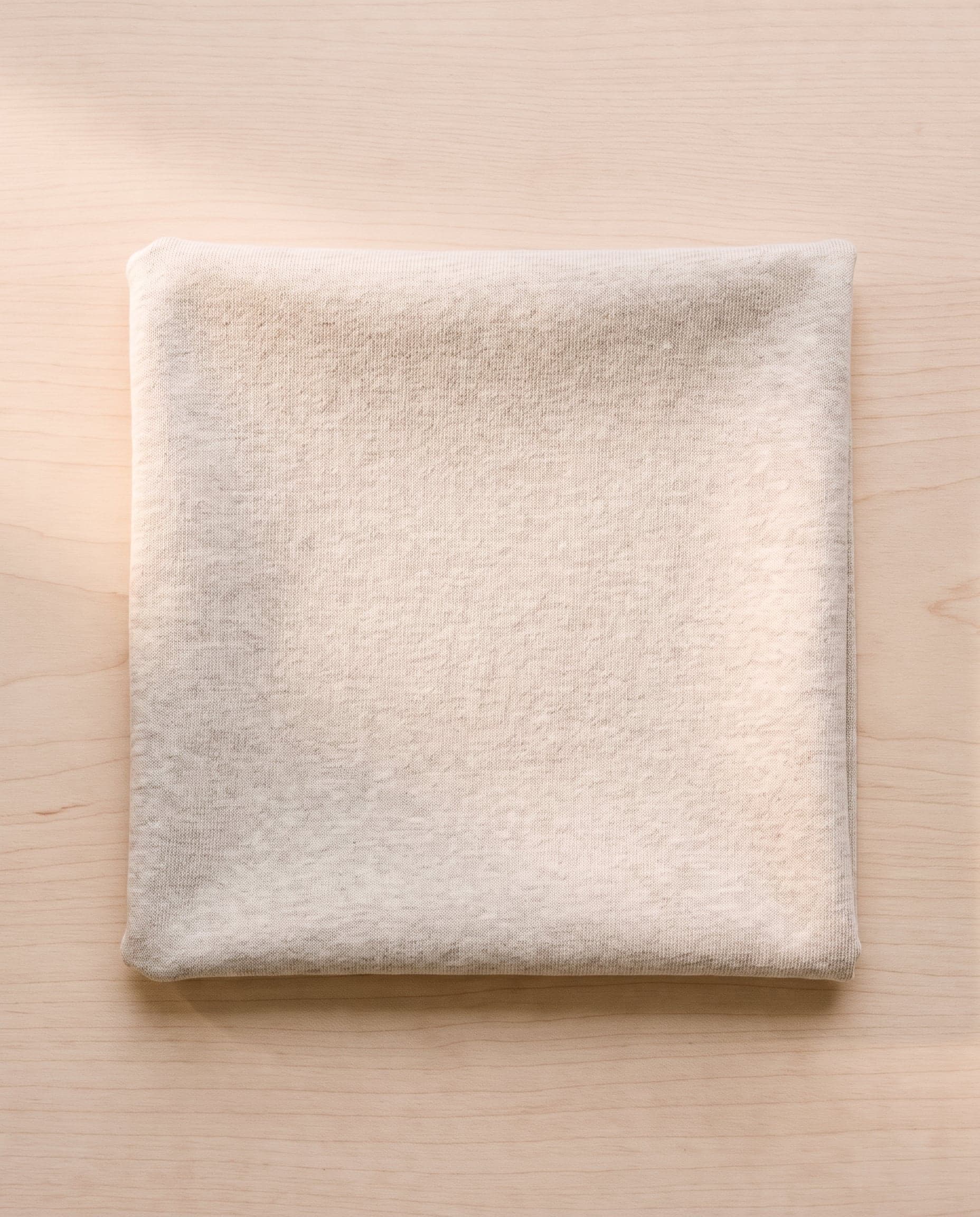 A folded square of oatmeal cotton jersey on pale ash wood.