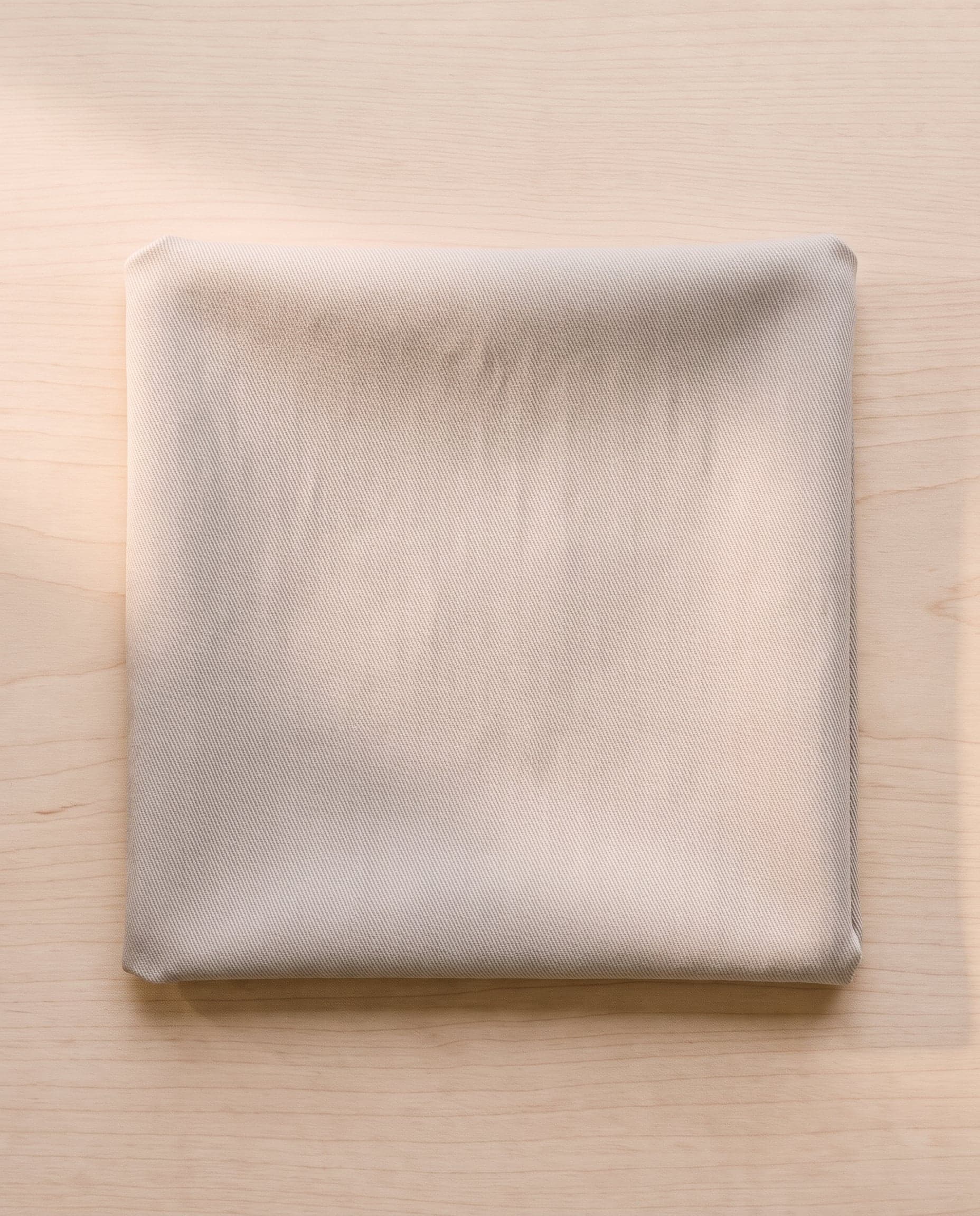 A folded square of stone cotton twill on pale ash wood.