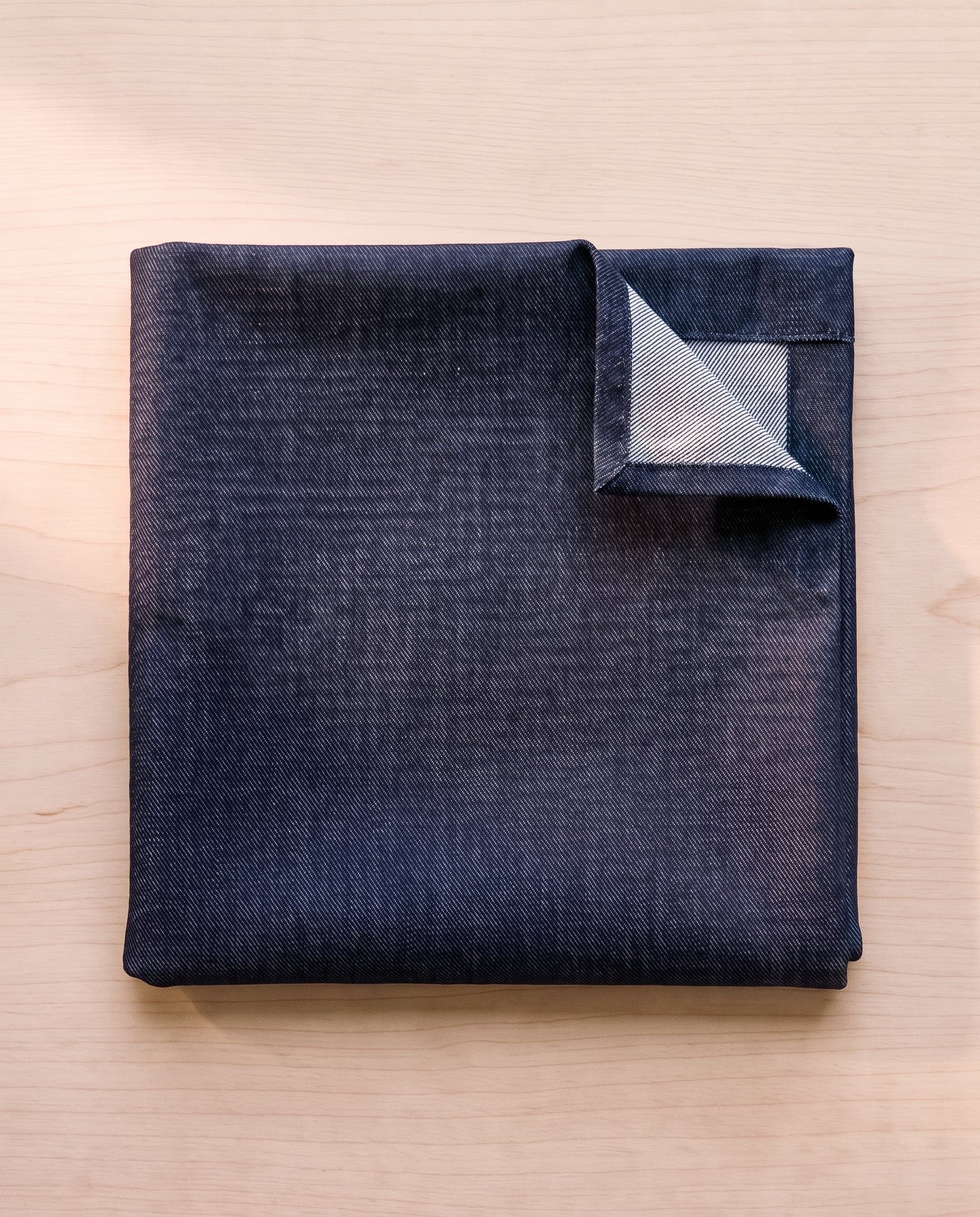 A folded square of indigo denim on pale ash wood, one corner turned back.