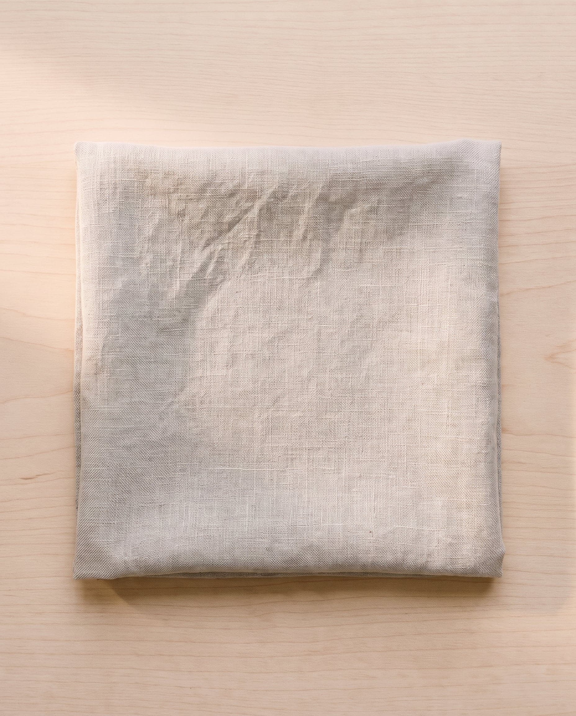 A folded square of natural flax linen on pale ash wood.